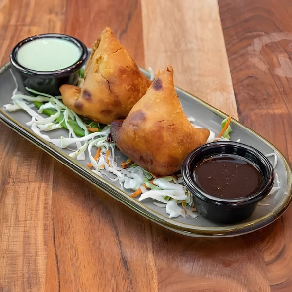 Two golden-brown samosas with shredded cabbage and carrots, served with green and brown dipping sauces.