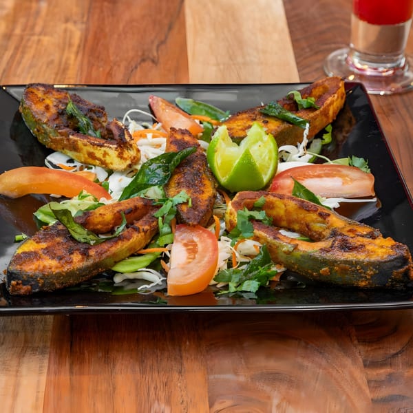 Masthi Indian Cuisine's fried fish, garnished with lime, tomatoes, and fresh herbs on a black plate.