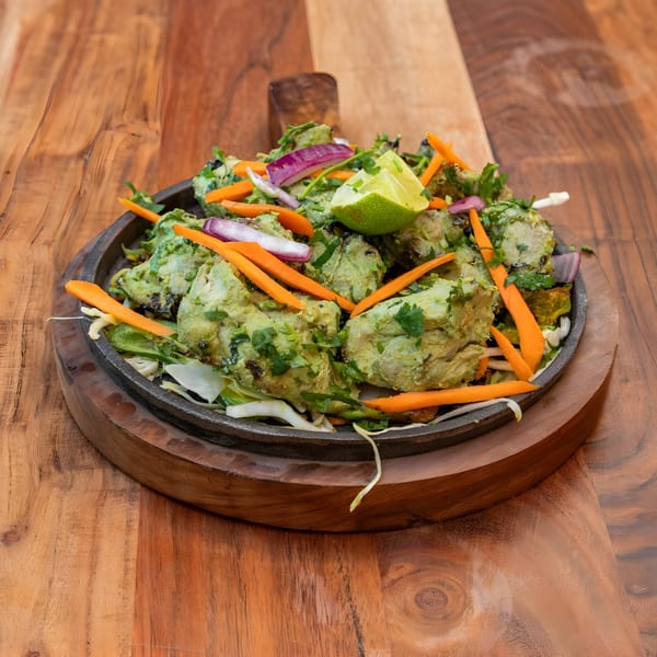 Masthi Indian Cuisine's green chicken tikka, garnished with carrots, red onion, and lime, on a wooden board.